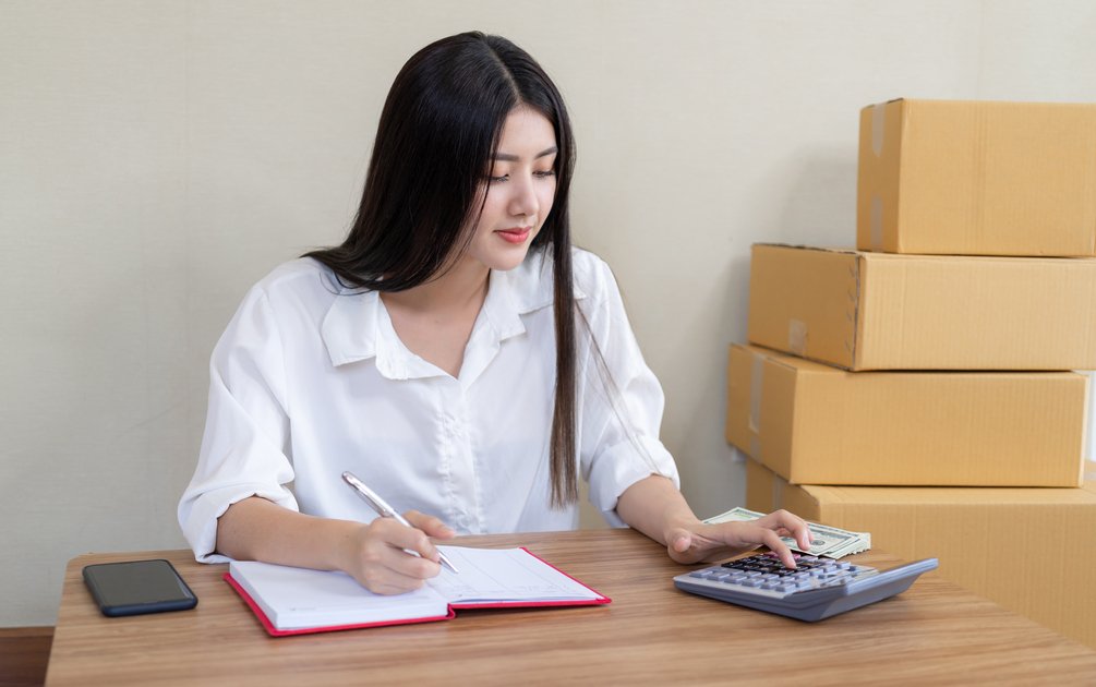 Wanita menghitung biaya bangun rumah menggunakan kalkulator dan mencatat anggaran di meja