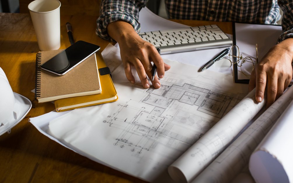 Arsitek sedang menganalisis gambar denah rumah klasik di atas meja kerja dengan dokumen perencanaan.