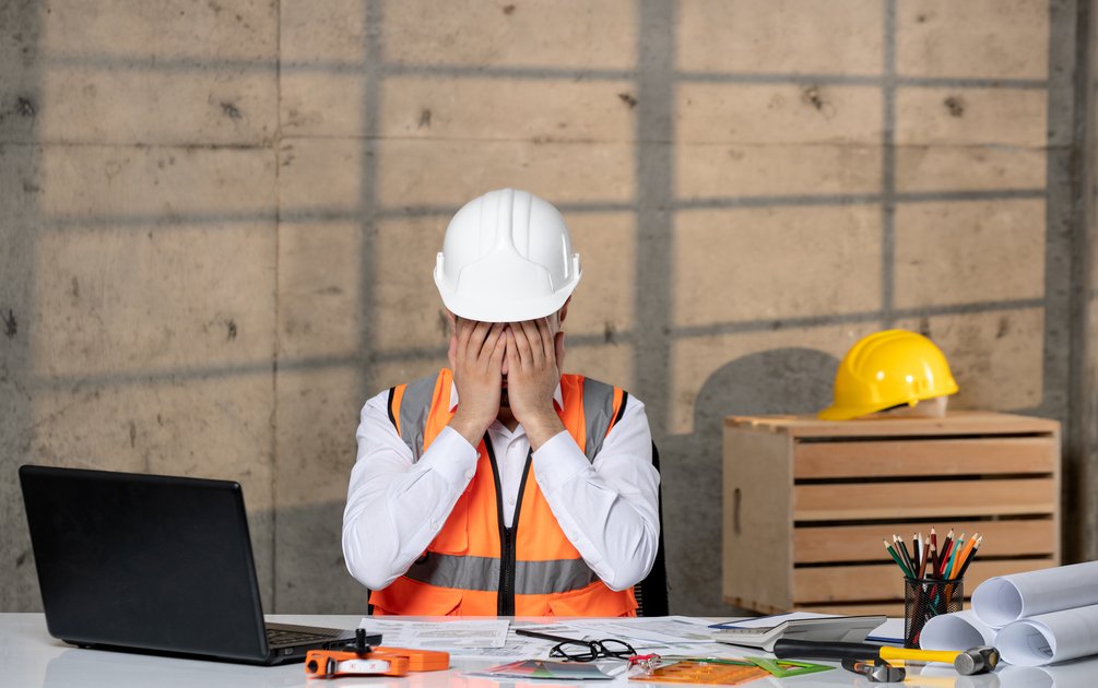 Arsitek muda tampak frustrasi di meja kerja dengan dokumen gambar bangunan, menggambarkan risiko kesalahan desain rumah yang bisa dihindari