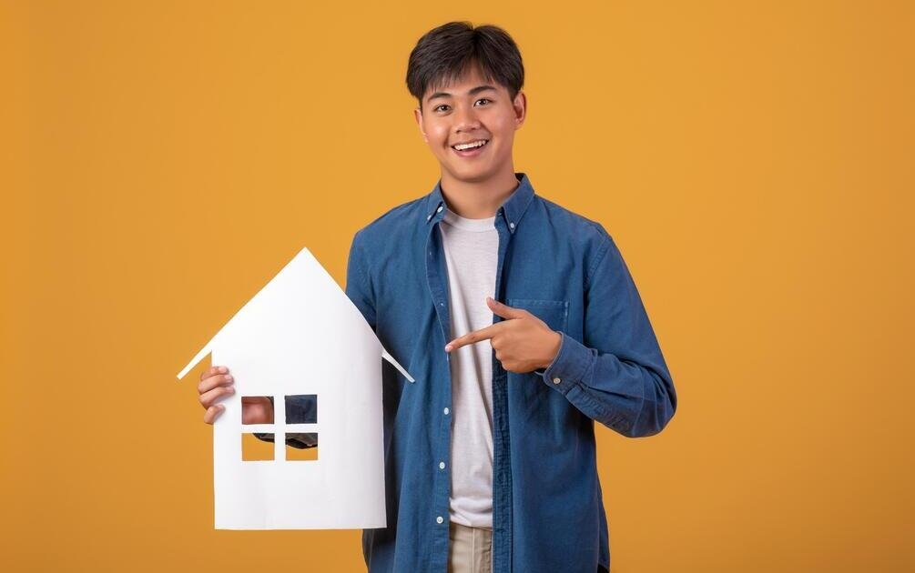 Pria muda memegang karton berbentuk rumah, menggambarkan impian membangun rumah dan pertimbangan menggunakan jasa arsitek