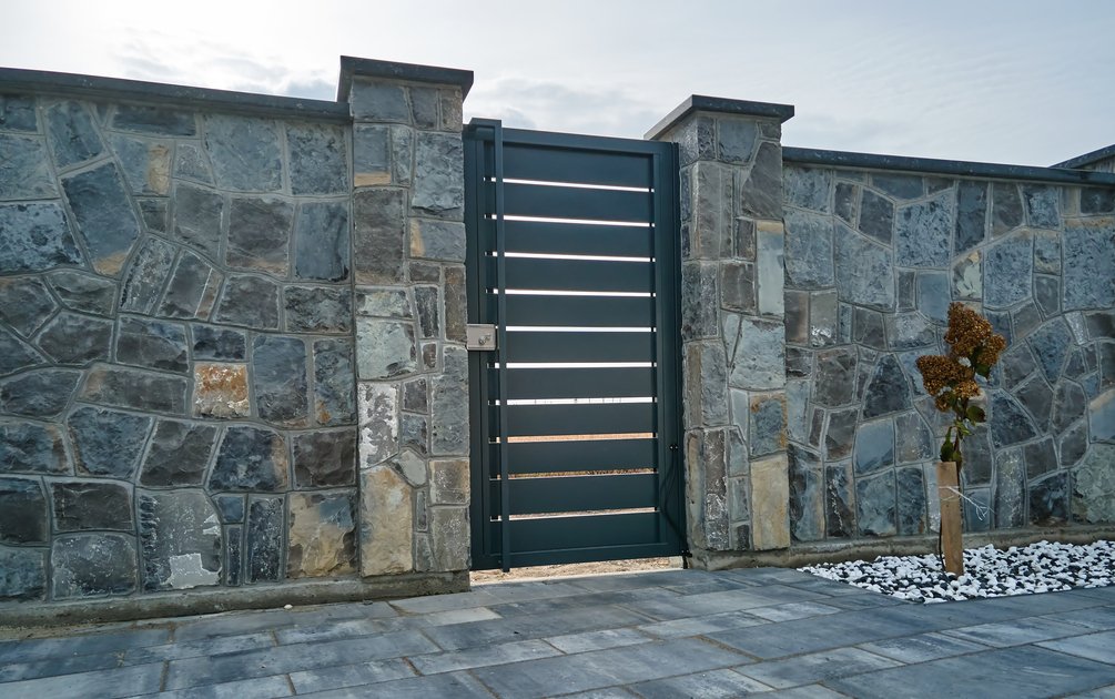 Pagar rumah batu alam gaya modern dengan pintu besi berwarna anthracite dan halaman berbatu — contoh desain fasad hunian yang kuat dan elegan