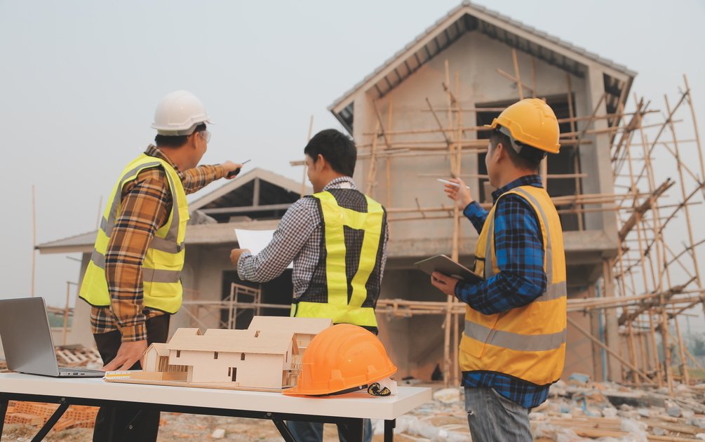 Tim arsitek jasa desain rumah Bogor sedang berdiskusi di lokasi proyek dengan maket rumah, gambar kerja, dan tablet untuk koordinasi konstruksi