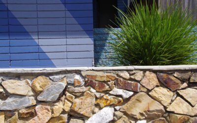 Pagar batu alam dengan kombinasi warna kuning, oranye, dan krem pada rumah modern tropis dengan tanaman ornamental