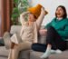 Dua perempuan tertawa bersama di ruang tamu dengan aksen bantal kuning hangat, menggambarkan psikologi warna ruang tamu yang mendorong interaksi