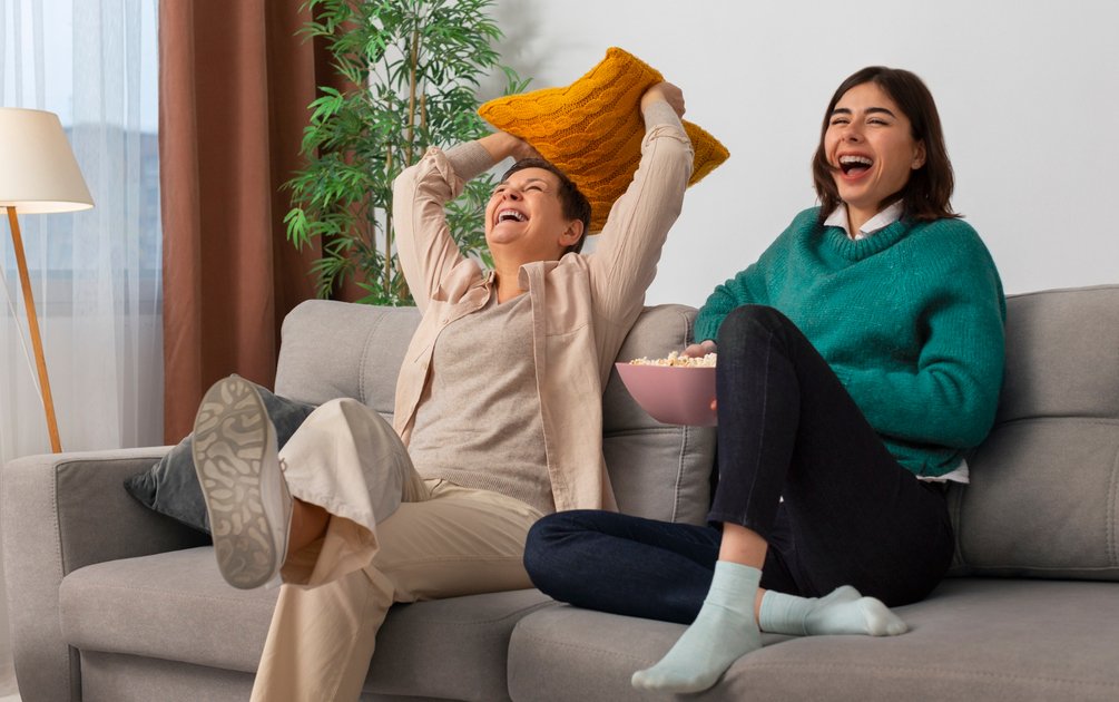 Dua perempuan tertawa bersama di ruang tamu dengan aksen bantal kuning hangat, menggambarkan psikologi warna ruang tamu yang mendorong interaksi