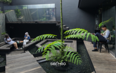 Courtyard tropis di kompleks Sibambo Studio HQ dan IDROC Space dengan pohon paku tanaman, deck kayu, dan pengunjung yang menikmati ruang