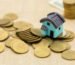 Miniatur rumah biru di atas tumpukan koin emas, ilustrasi pentingnya menghitung biaya jasa arsitek sejak awal perencanaan budget rumah