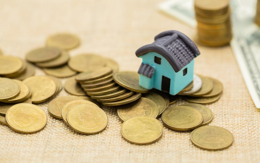 Miniatur rumah biru di atas tumpukan koin emas, ilustrasi pentingnya menghitung biaya jasa arsitek sejak awal perencanaan budget rumah