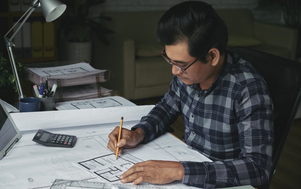 Arsitek profesional mengerjakan denah rumah dengan kalkulator dan referensi, ilustrasi cara memilih jasa arsitek rumah yang tepat